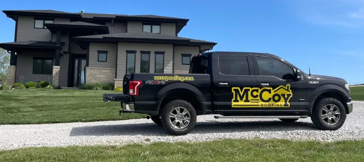 A black pickup truck with "McCoy Roofing" branding is parked on a gravel driveway in front of a modern two-story house.