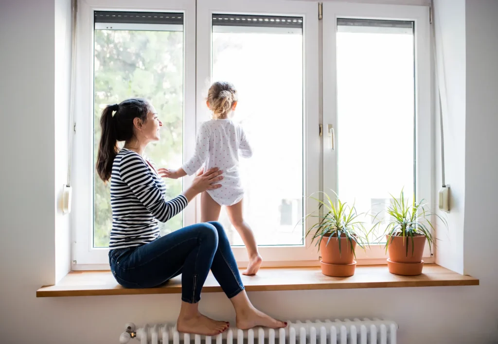 A woman sits on a windowsill watching a young child standing and looking outside. Two potted plants are on the windowsill.
