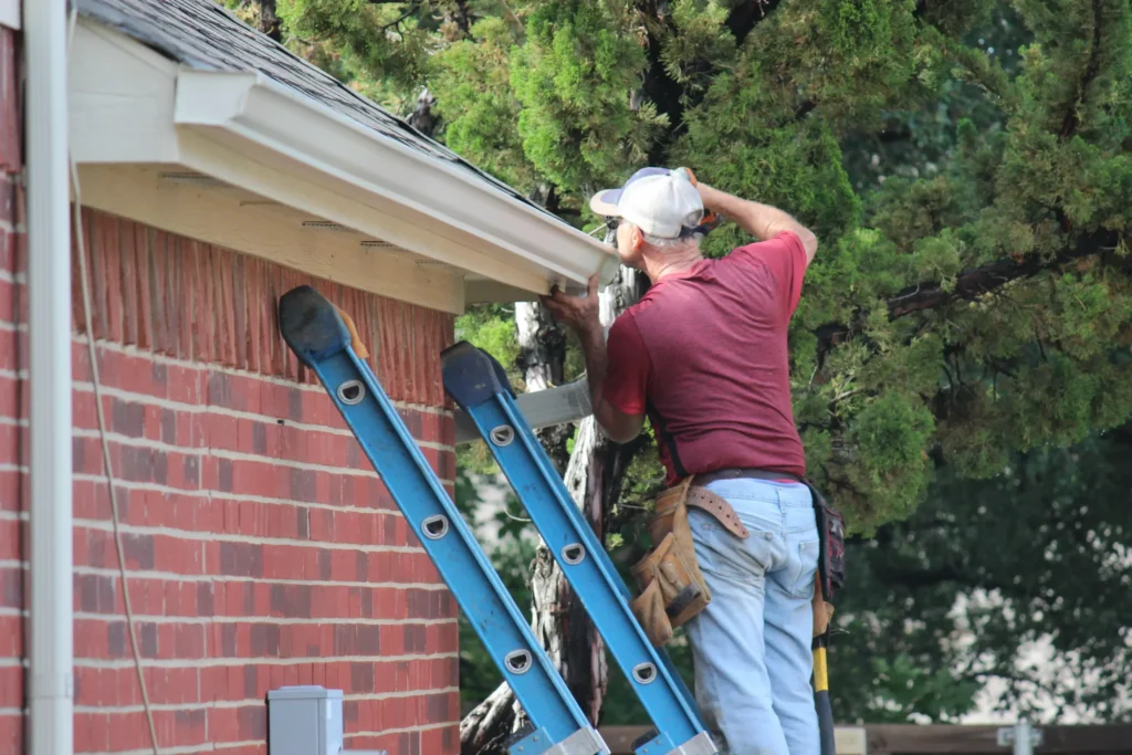 A man in a maroon shirt and jeans stands on a ladder, working on the gutter of a brick house next to a green tree.