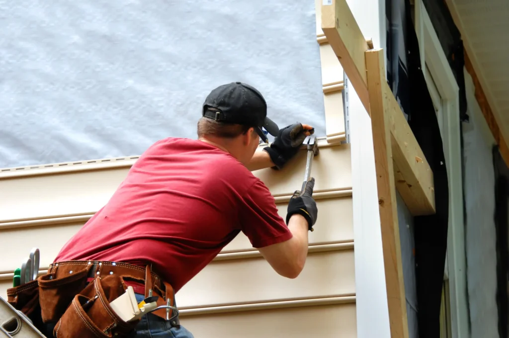 A worker in a red shirt installs beige vinyl siding on a house, using a hammer and nails. A tool belt with various tools is strapped around their waist.